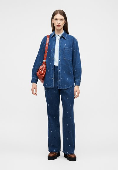Femme portant une chemise en jean bleue assortie à un pantalon avec de petits motifs blancs, des chaussures compensées marron et un sac à bandoulière rouge.