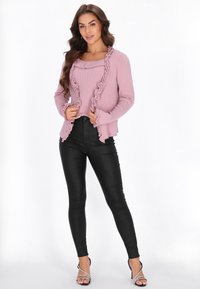 Różowy kardigan z prążkowaną teksturą i falbanami, noszony na dopasowanej bluzce, w zestawie z czarnymi legginsami z sztucznej skóry i sandałkami na obcasie.