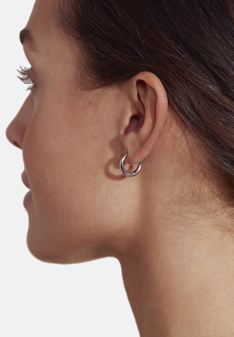 Silver hoop earring worn on the left ear, featuring a round shape and smooth texture. The design is simple and modern, reflecting a minimalist style.