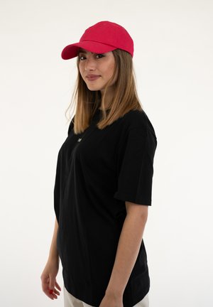 Tricou oversized negru, cu mâneci scurte; asortat cu o șapcă roz aprins. Tricoul are un mic logo pe față. Textură de țesătură netedă.