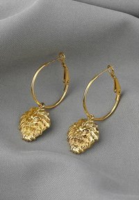 Goldene Ohrstecker mit detaillierten Löwenkopf-Anhängern, polierter Oberfläche und Federkopfverschluss, vor einem strukturierten grauen Stoffhintergrund.