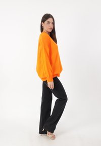 Maglione arancione oversize a maglia con polsini a costine, abbinato a jeans neri svasati e sandali color crema, su uno sfondo bianco.