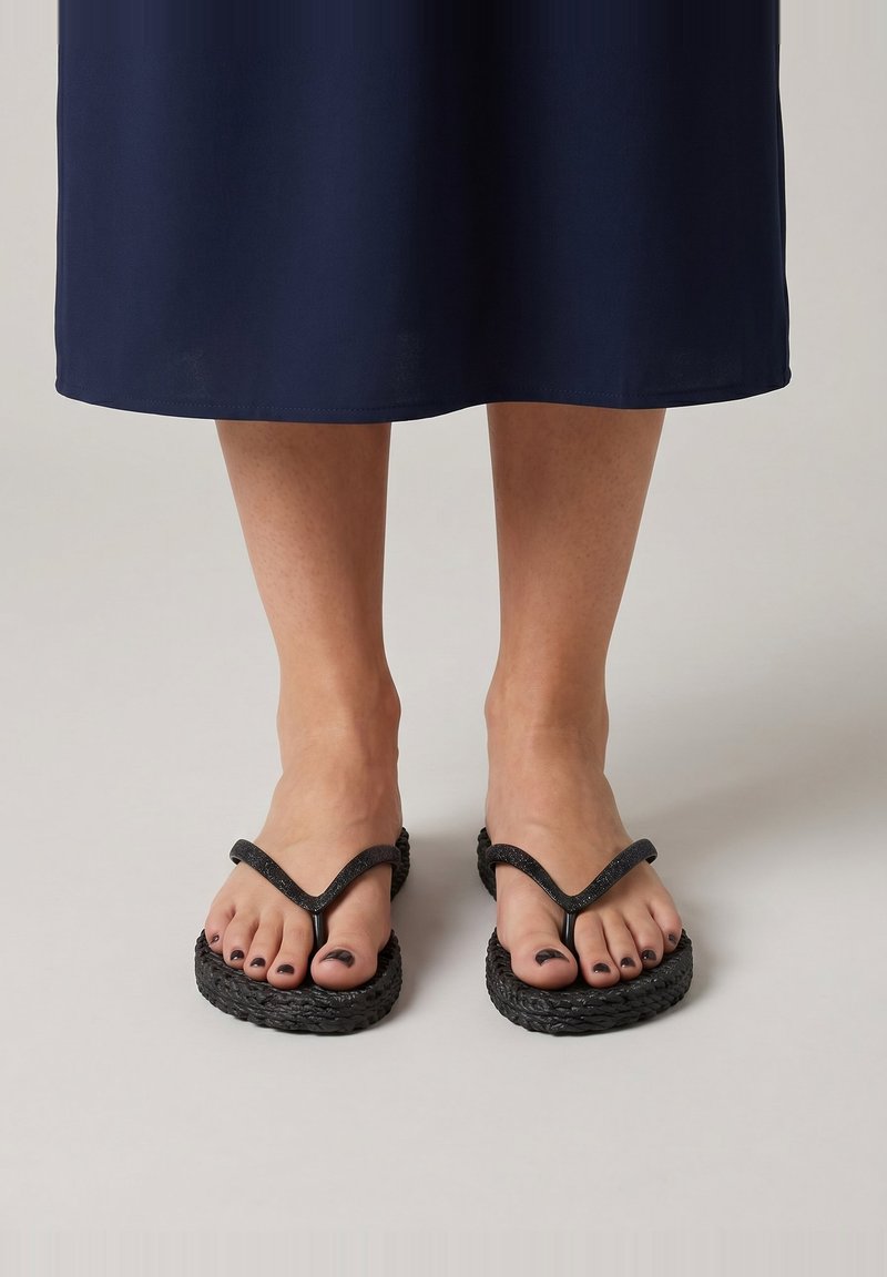 Pies con esmalte de uñas oscuro que llevan chanclas de plataforma tejidas negras y una falda azul marino visible hasta la mitad de la pantorrilla sobre un fondo liso.