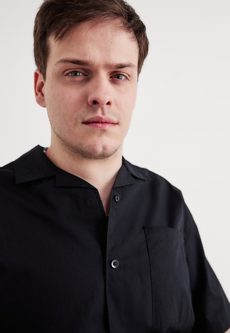 Svart kortermet skjorte med knapper, en brystlomme, krage og glatt tekstur. Stoffet virker lett og matte.