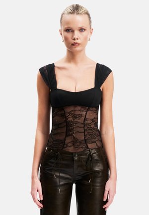Vrouw met blond haar in een zwart kanten korset top en zwarte leren broek staat tegen een effen witte achtergrond.