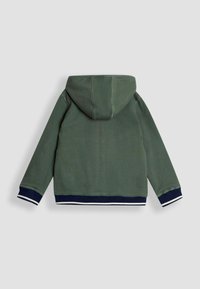 Tumši zaļš korduroja kapuce ar ribotu tekstūru, ar tumši zilu piedurkņu un apakšēju malu apdari baltā krāsā. Aizmugures skats, priekšpuses detaļas nav redzamas.