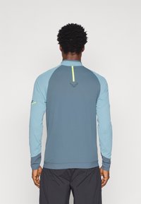 Tricou sportiv cu mâneci lungi, de culoare albastră, cu un panou din spate de un albastru mai închis. Prezintă un accent vertical galben și detalii cu logo aproape de decolteu.