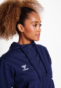Marineblauwe zip-up hoodie met een hoge kraag, voorzien van een wit logo op de borst, met een gestructureerde stof en verstelbare koordjes bij de capuchon.
