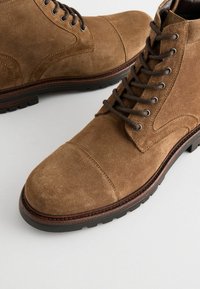 Botine din suede maro, cu vârful rotund, dotate cu șireturi întunecate și talpă din cauciuc negru. Cusături contrastante pe margine.