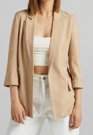 Femme portant un blazer beige avec les manches retroussées, un crop top blanc et un pantalon blanc taille haute sur un fond uni.