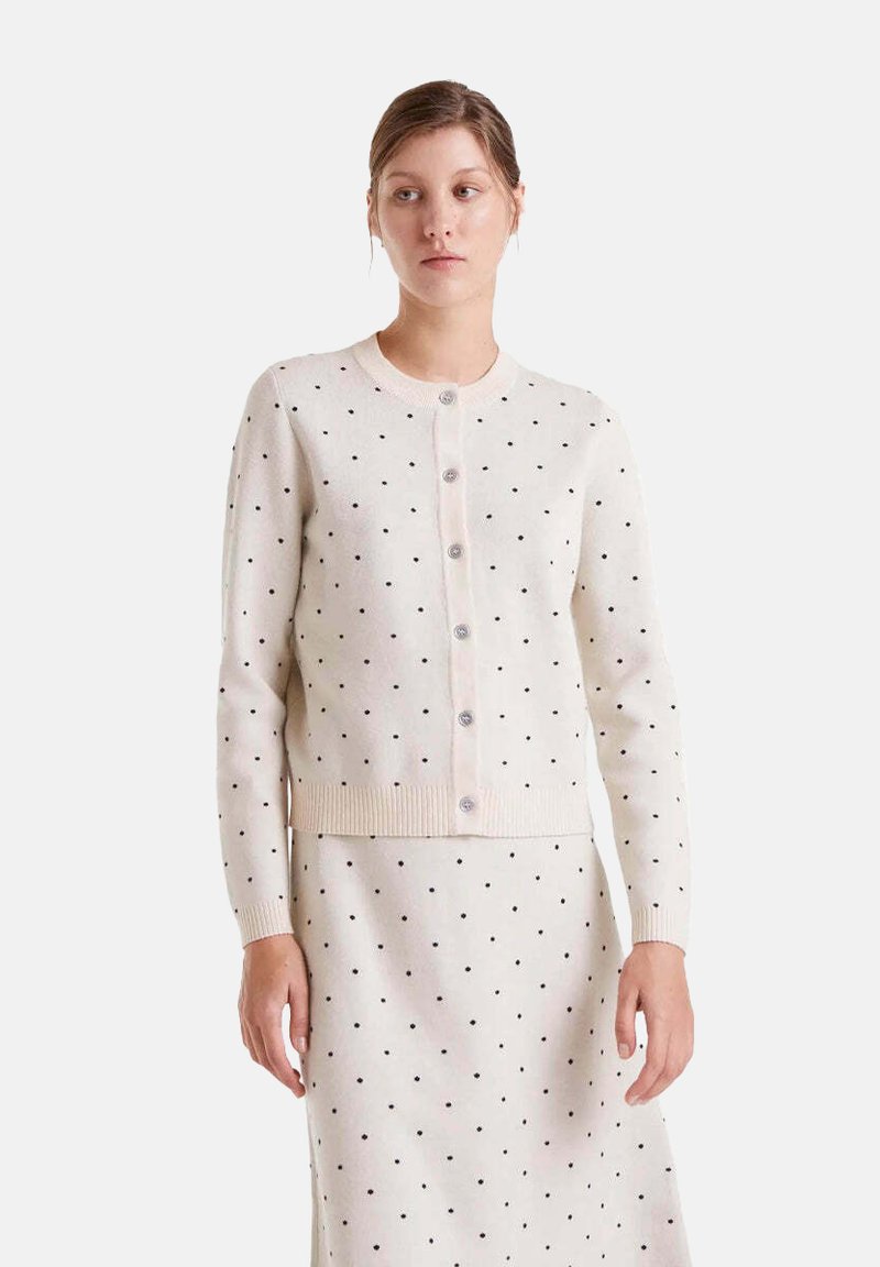 Mujer vestida con un cárdigan de botones color crema y una falda a juego con pequeños lunares negros, de pie frente a un fondo liso.