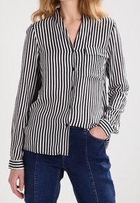 Camisa de manga larga a rayas blancas y negras con cierre de botones, cuello y bolsillo. Se lleva con vaqueros azul oscuro. Textura de tela lisa.