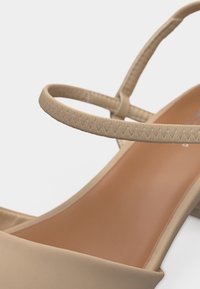 Sandale beige pour femme avec une fine bride de cheville cousue et un design à bout ouvert sur un fond clair.