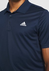 Námořnická modrá polo tričko s límcem, na levé straně obsahující bílé logo Adidas, nošené osobou od hrudníku nahoru.