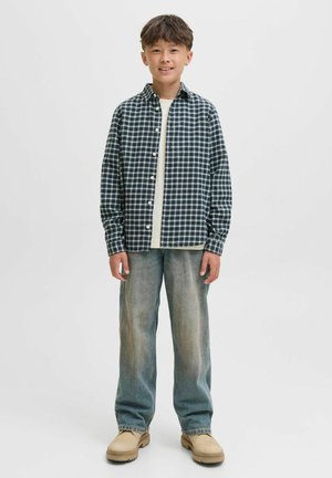 Chemise à carreaux bleu marine et blanche, superposée à un T-shirt beige. Jean en denim clair ample et chaussures beiges.