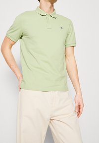 Homme portant un polo à manches courtes vert clair et un pantalon beige, une main posée sur la hanche, se tenant devant un fond uni.