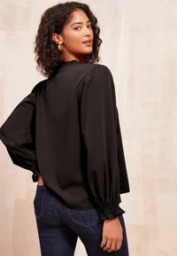 Black blouse with a high neck, puffed sleeves, and ruffled trim. Textured fabric, loose fit, and elastic cuffs. Paired with dark jeans.