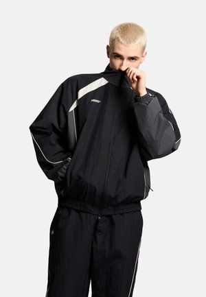 Młoda osoba z krótkimi blond włosami w czarno-szarym dresie, częściowo zakrywająca twarz kołnierzem kurtki na jednolitym tle.