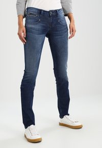 Mörkblå jeans med slim fit, med framfickor och dragkedjedetalj. Kombinationen med vita sneakers och en topp med grå ärmar.