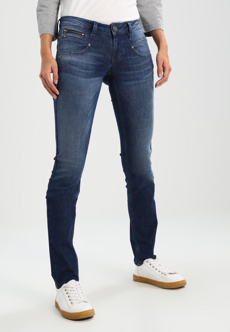 Mörkblå jeans med slim fit, med framfickor och dragkedjedetalj. Kombinationen med vita sneakers och en topp med grå ärmar.