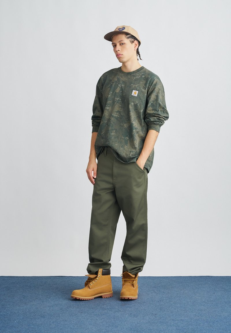 Young man with braided hair wears green camo sweatshirt, olive pants, tan work boots, and beige cap, standing on blue carpet against white wall.