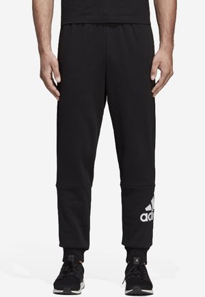 Mand iført sorte Adidas joggingbukser med hvidt logo på venstre læg og sorte sneakers, stående med armene afslappet ned langs siderne.