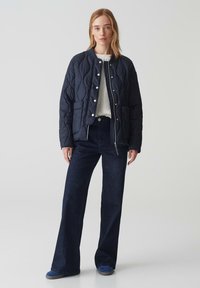 Giacca trapuntata blu navy con bottoni a pressione, tasche laterali, indossata sopra un maglione crema e jeans neri a zampa, abbinata a scarpe blu.