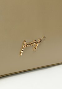 Złote logo na gładkim, matowym khaki tle, z subtelnym połyskiem. Logo ma kursywny design z płynącymi liniami.