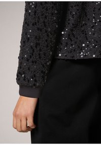 Top negro con lentejuelas y tela texturizada, presenta un puño acanalado y un ajuste relajado. Los detalles incluyen lentejuelas brillantes de diferentes tamaños.