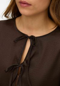 Smeđa bluzica teksture s okruglim izrezom, isječkom u obliku ključa sa vezanjem i lagano drapiranom tkaninom.