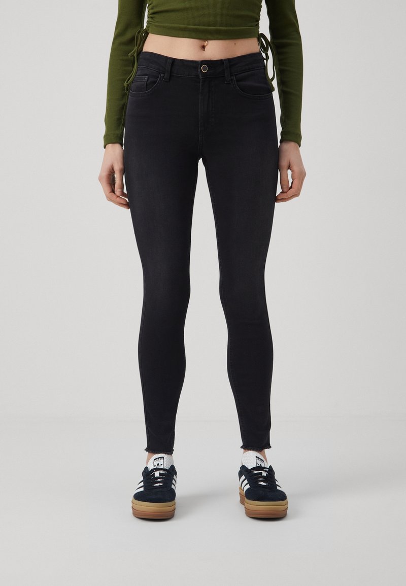 Femme portant un jean skinny noir taille haute, un top vert à manches longues court et des baskets Adidas noires avec des rayures blanches sur un fond uni.