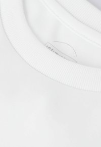 Bílá bavlněná tričko s žebrovaným kulatým výstřihem. Obsahuje malou nálepku uvnitř límce. Hladká textura, minimalistický design.