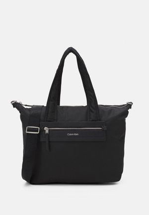 Sac cabas noir Calvin Klein avec double poignée, bandoulière ajustable et poche zippée à l'avant sur fond uni.
