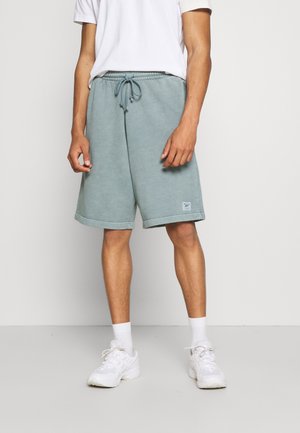 Hombre vestido con pantalones cortos de cordón color verde azulado claro, zapatillas blancas, calcetines blancos y una camiseta blanca, posando frente a un fondo liso.