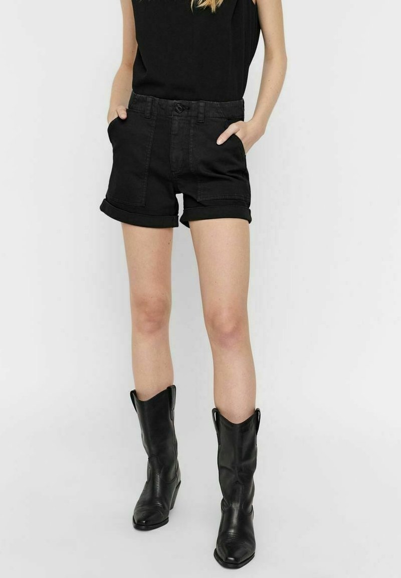 Personne portant un haut noir sans manches, un short noir retroussé avec les mains dans les poches, et des bottes de cowboy en cuir noir mi-mollet sur fond blanc.