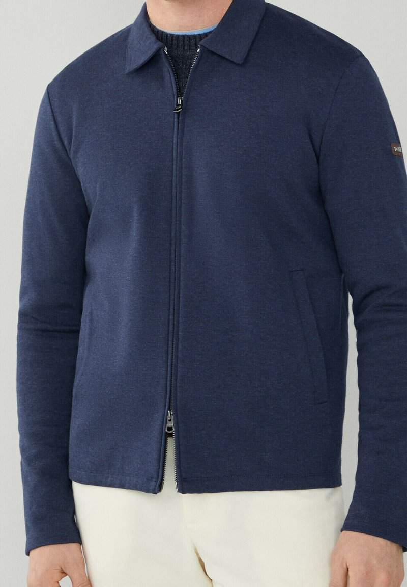 Homme portant une veste zippée bleu marine sur un pull noir et un pantalon beige clair sur un fond uni.