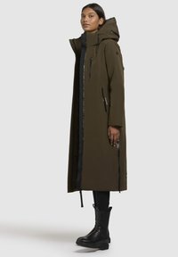 Lång olivgrön parka med huva, med svarta dragkedjor, en oversized design och ett slätt, vattenavvisande tyg.