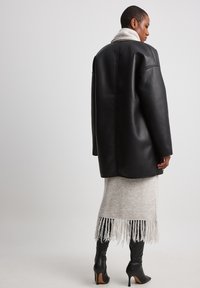 Giacca oversize nera in pelle con dettagli nelle cuciture, abbinata a una gonna in maglia frangiata grigia chiara e stivali neri con tacco.