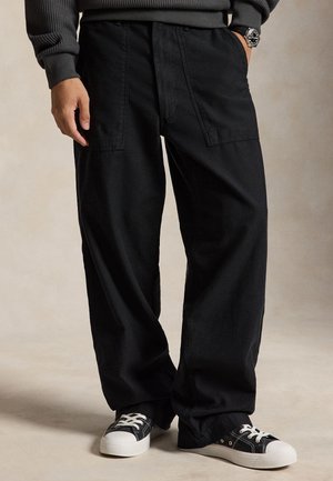 Personne portant un pantalon noir ample avec de grandes poches avant et des baskets basses noires avec semelles et lacets blancs, debout sur un fond neutre.