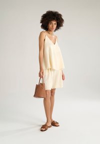 Top in seta gialla chiara con design a strati, abbinato a pantaloni corti coordinati. Borsa marrone con motivo in rilievo e sandali piatti marroni.