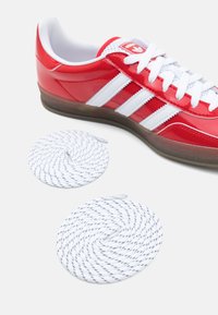 Red glossy sneaker with white stripes, white laces, and a textured brown sole. Includes two coiled white shoelaces with dark accents.