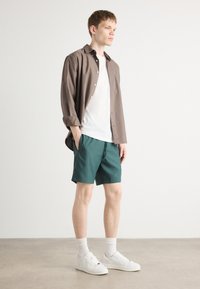 Tenue décontractée comprenant une chemise brune à boutons, un t-shirt blanc, un short turquoise, des baskets blanches et des chaussettes blanches de longueur mi-mollet. Textures lisses tout au long.