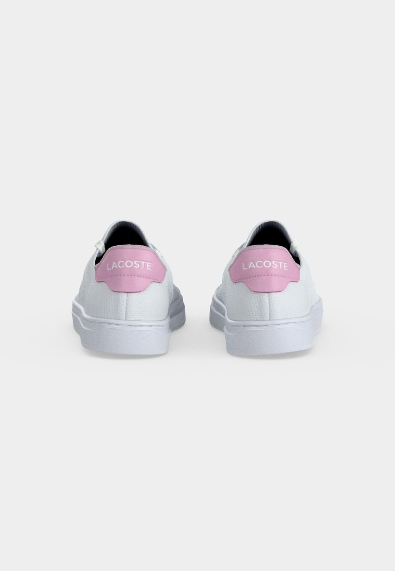 Weiße Low-Top-Sneaker mit pinkfarbenen Fersenlaschen mit der Aufschrift „Lacoste“, von hinten auf einem schlichten hellgrauen Hintergrund gezeigt.