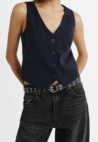 Marineblaues Weste mit tiefem V-Ausschnitt, fünf Knöpfen und zwei vorderen Taschen, kombiniert mit schwarzen, hochgewaisteten Jeans und einem genoppten Gürtel.