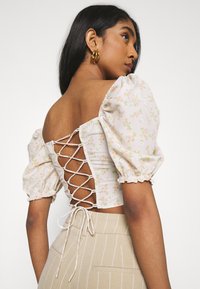 Blusa de manga curta com estampado floral em rosa claro, com mangas bufantes, cordões nas costas e detalhe de laço na cintura, combinada com calças bege listradas.