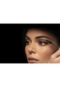 Kvinde påfører sort mascara på øverste øjenvipper, nærbillede af ansigt mod en sort baggrund.