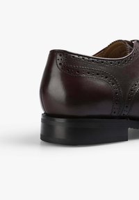 Zapato de cuero color burdeos oscuro con perforaciones decorativas a lo largo de las costuras, tacón bajo apilado y textura lisa.