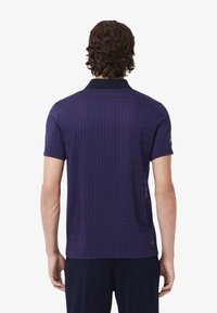 Kortärmad polo tröja i mörklila med ett strukturerat rutmönster och en kontrasterande svart krage. Tillverkad av andningsbart material för komfort.