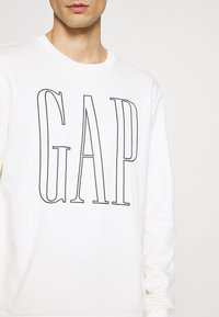 Felpa bianca con logo "GAP" in outline nero sul davanti. Presenta un colletto rotondo e maniche lunghe. Tessuto morbido e testurizzato.
