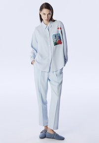 Camicia azzurra con bottoni, decorata con una patch floreale sul petto, abbinata a pantaloni azzurri e scarpe blu coordinate. Tessuto morbido.